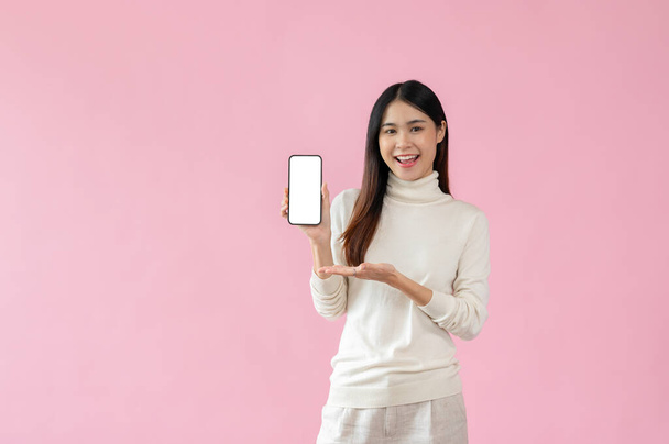 Veselý a krásný asijské žena v ležérní oblečení je zobrazování smartphone bílá obrazovka mockup na kameru, zatímco stojí proti izolované růžové pozadí studia. - Fotografie, Obrázek