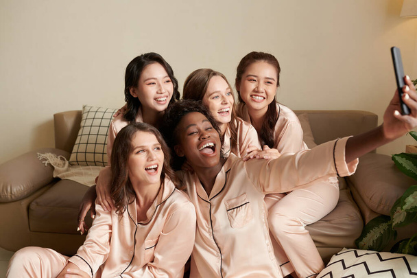 Radosne koleżanki w satynowych piżamach robiące selfie na smartfonie - Zdjęcie, obraz