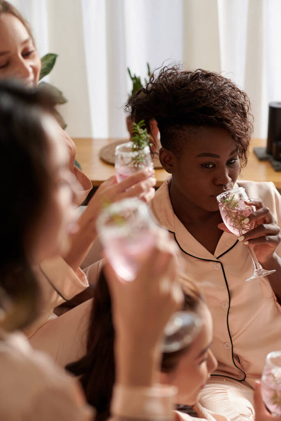 Pensive jovem mulher negra bebendo espumante vinho rosa de óculos decorados ao ter dormida com amigos - Foto, Imagem