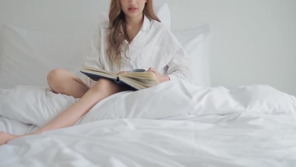 Νεαρή γυναίκα πίνει καφέ ενώ διαβάζει ένα βιβλίο που κάθεται στο κρεβάτι το πρωί. - Πλάνα, βίντεο