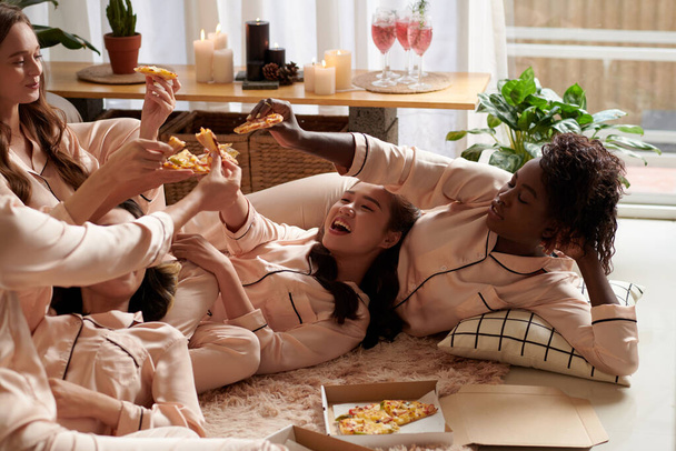 Группа молодых женщин тосты с ломтиками пиццы, когда они лежат на полу в пижаме - Фото, изображение