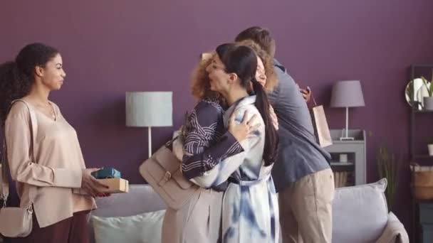Cinco jóvenes amigos multiétnicos se saludan y se abrazan cuando se reúnen para cenar en un apartamento elegante, intercambiando regalos - Imágenes, Vídeo