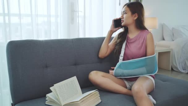 Mladá asijská žena nošení měkké dlahy kvůli bolavé rameno pomocí telefonu na pohovce doma. - Záběry, video