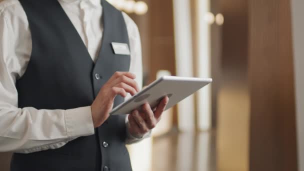 Sezione media di operaio alberghiero irriconoscibile che indossa uniforme in piedi nella hall o nella lounge utilizzando tablet digitale - Filmati, video