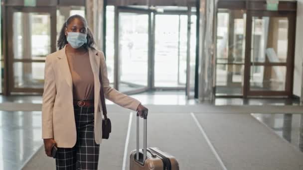 Retrato médio longo da jovem mulher negra vestindo roupas elegantes com máscara protetora em pé com mala no lobby do hotel - Filmagem, Vídeo