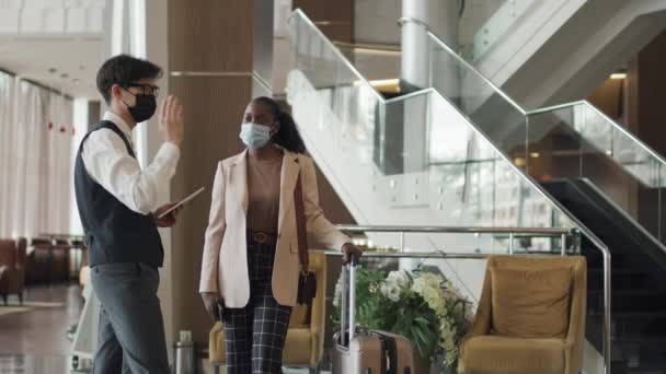 Jovem adulto asiático vestindo uniforme e máscara protetora trabalhando com hóspedes afro-americanos em hotel moderno - Filmagem, Vídeo