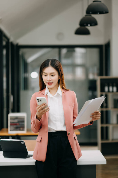 Confident Asian woman with a smile standing holding smartphone at the modern office - Fotó, kép
