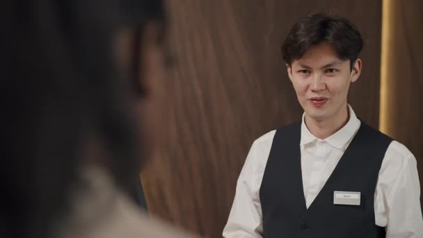 Nuoren aasialaisen vastaanottovirkailijan olkapään yli valikoiva tarkennus, joka työskentelee uuden hotellin vieraan kanssa - Materiaali, video