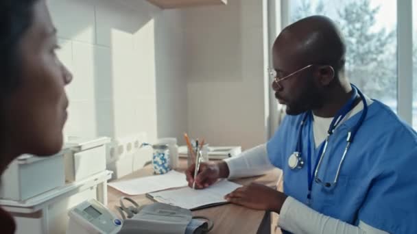 Medico afroamericano seduto alla scrivania a prendere appunti nella scheda medica mentre conversava con una paziente donna - Filmati, video