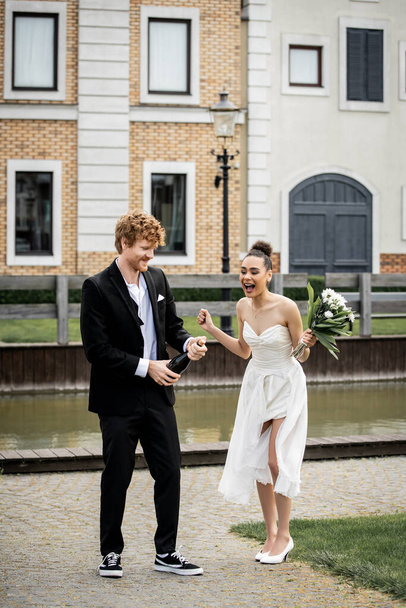 young redhead groom opening champagne near african american bride with bouquet on street - Φωτογραφία, εικόνα