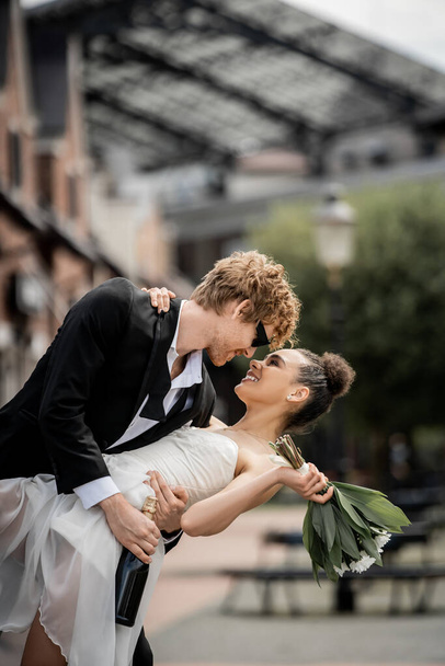 stylish redhead groom embracing african american bride wit bouquet on city street, outdoor wedding - Photo, Image