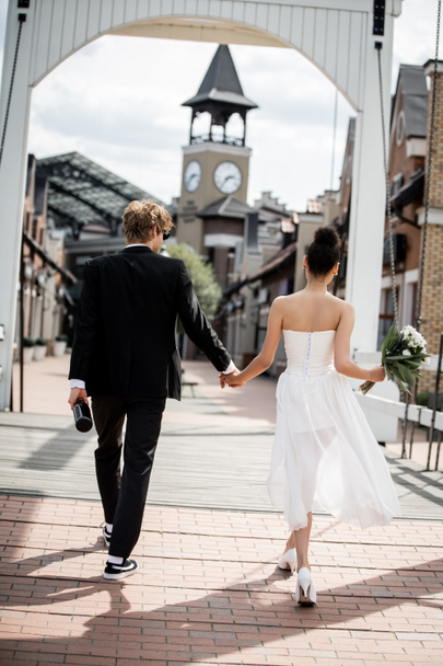 back view of multiethnic couple with champagne and flowers holding hands on bridge, wedding outdoors - Photo, Image