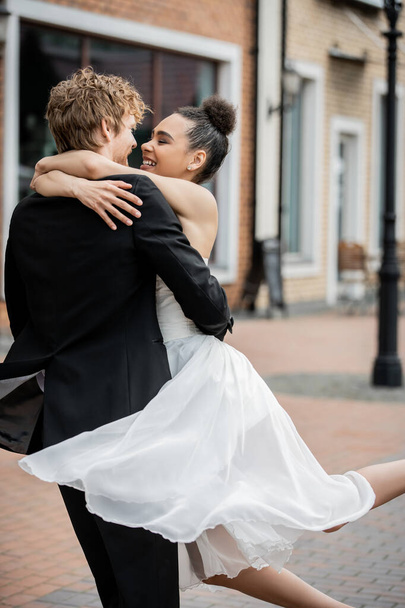 ceremonia de boda al aire libre, pareja interracial encantado y elegante abrazando en la calle de la ciudad - Foto, imagen