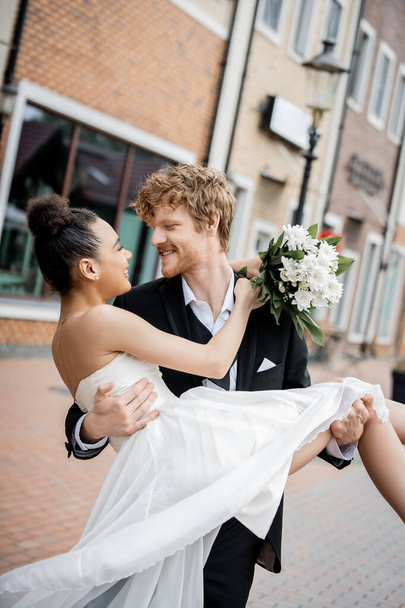 urban romance, redhead groom holding african american bride with wedding bouquet on street - Photo, Image