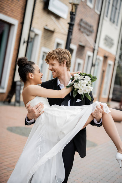 love in city, happy young groom holding african american bride with flowers on street - Foto, Imagem