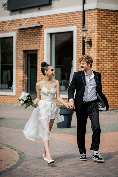 elegant multiethnic couple in wedding attire holding hands and walking on urban street - Foto, afbeelding