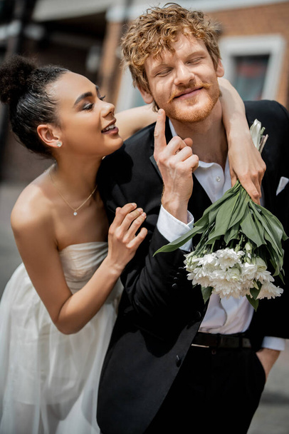 šťastný ryšavý muž ukazuje na tvář blízko šťastný africký americký nevěsta s květinami, svatba venku - Fotografie, Obrázek