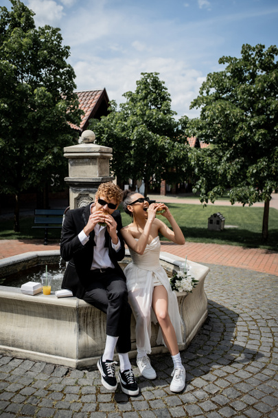 элегантные межрасовые молодожены в солнечных очках едят бургеры возле фонтана в европейском городе - Фото, изображение
