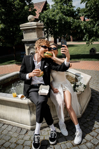african american woman with burger feeding groom taking selfie on smartphone near city fountain - Photo, Image