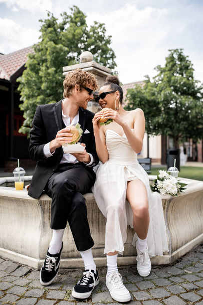 radostné mezirasové novomanželé ve slunečních brýlích drží hamburgery a dívají se na sebe blízko fontány - Fotografie, Obrázek