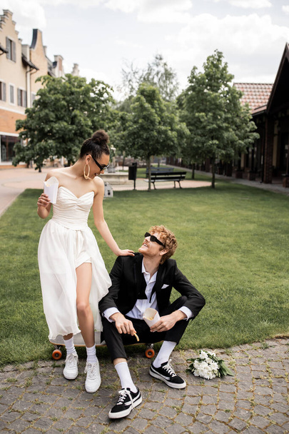 redhead groom sitting on longboard near african american bride with french fries, outdoor wedding - Foto, afbeelding