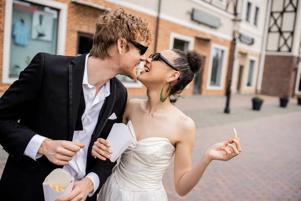 urban romance, outdoor wedding, multiethnic newlyweds in sunglasses eating french fries together - Photo, Image
