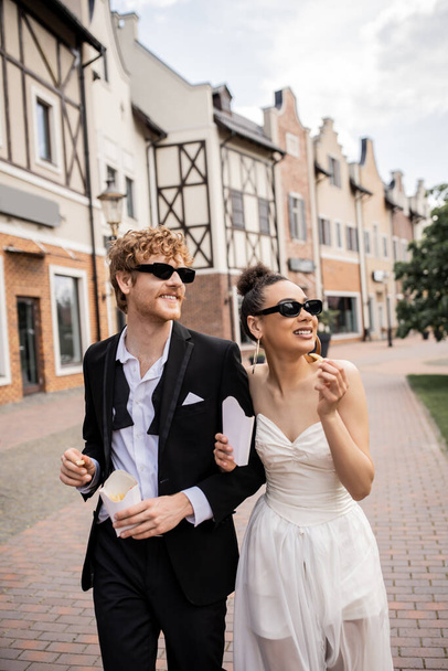 stylish multiethnic couple with french fries walking in city, wedding attire, sunglasses, happiness - Фото, зображення