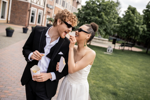 happy african american bride feeding stylish groom with french fries, wedding on city street - Photo, Image