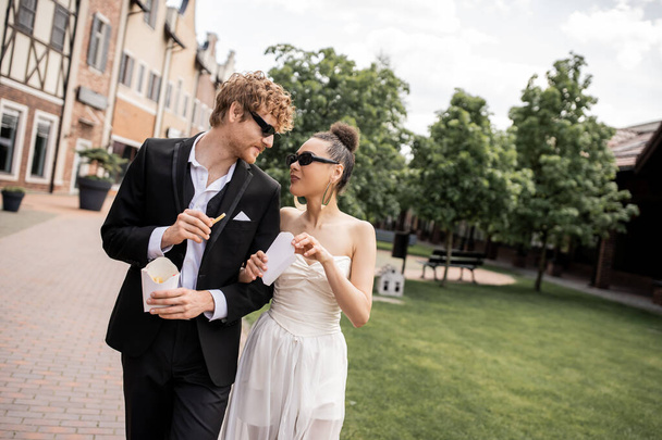 interracial newlywed couple in sunglasses, with french fries, looking at each other on street - Photo, Image