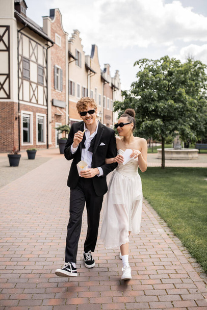 redhead man and african american woman walking with french fries in city, wedding attire, sunglasses - Zdjęcie, obraz