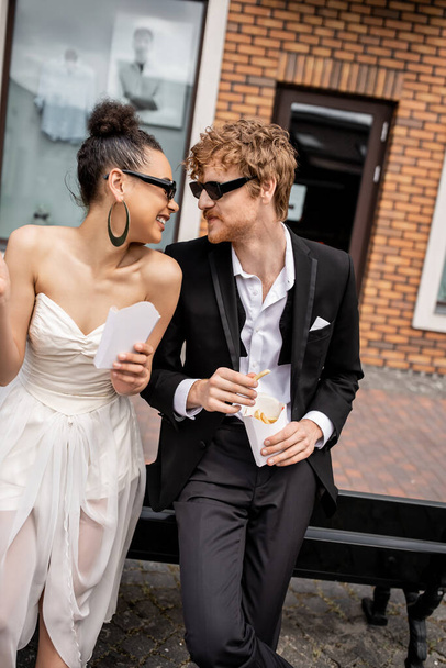 fröhliches multiethnisches Paar mit Sonnenbrille und Pommes, das sich auf der Straße anschaut - Foto, Bild