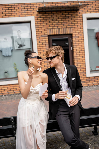 african american bride with french fries smiling near elegant redhead groom, wedding in modern city - 写真・画像