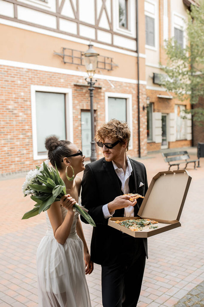 stílusos többnemzetiségű friss házasok a városi utcában, pizza virágokkal, esküvői ruhával, napszemüveggel - Fotó, kép