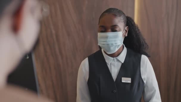 Selektivní zaměření přes rameno záběr nerozpoznatelné černošky žena nosí masku pracující na recepci hotelu mluvit s asijskými hosty - Záběry, video