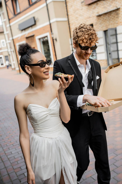 outdoor celebration, interracial couple walking with pizza in city, wedding attire, sunglasses - Foto, Bild