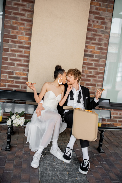 elegant multiracial couple with pizza kissing on bench, wedding in city, outdoor celebration - Photo, Image
