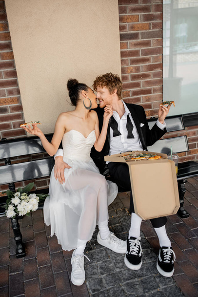 young interracial newlyweds with pizza sitting on bench in european city, outdoor celebration - Фото, зображення