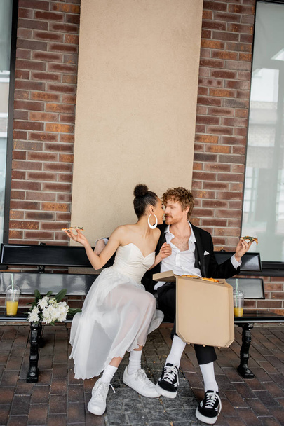 romantic interracial couple in wedding attire, with pizza, kissing on bench, celebration in city - Zdjęcie, obraz