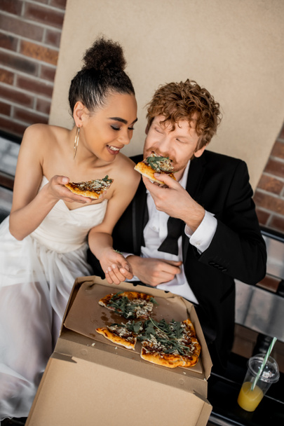 zadowolona wielonarodowa para w stroju ślubnym jedząca smaczną pizzę na ławce, ślub w mieście - Zdjęcie, obraz