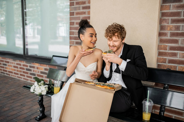 cheerful, elegant multiethnic newlyweds eating pizza near flowers and orange juice on bench in city - 写真・画像