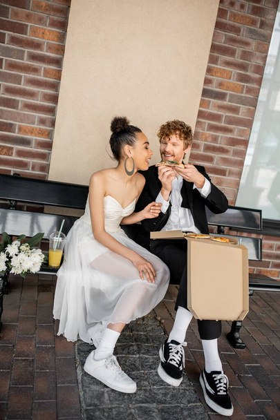 stylish and happy interracial newlyweds eating pizza near orange juice and flowers on bench in city - Photo, Image