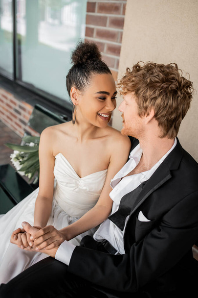 alegre pareja interracial cogidas de la mano y mirándose en el banco, boda en la ciudad - Foto, imagen