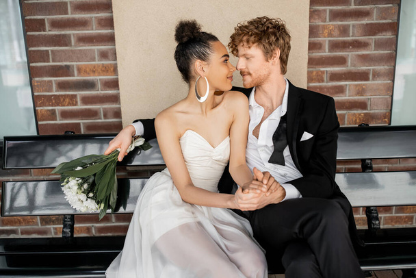 redhead man with flowers sitting face to face with african american bride on bench, outdoor wedding - Foto, imagen