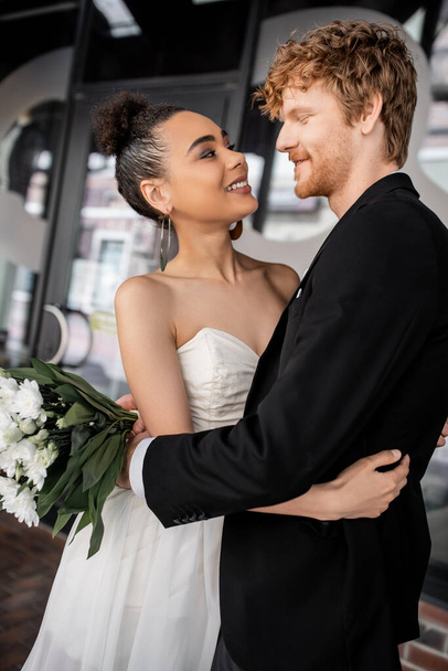 overjoyed and elegant interracial couple in wedding attire embracing on city street - 写真・画像