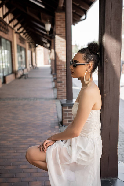 zijaanzicht van charmant Afrikaans amerikaanse vrouw in trouwjurk en zonnebril op stedelijke straat - Foto, afbeelding