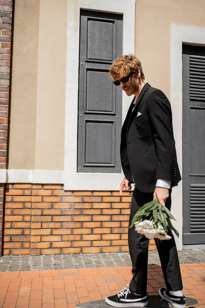 елегантний чоловік в чорному костюмі і сонцезахисних окулярах, що йдуть по міській вулиці, наречений з весільним букетом - Фото, зображення