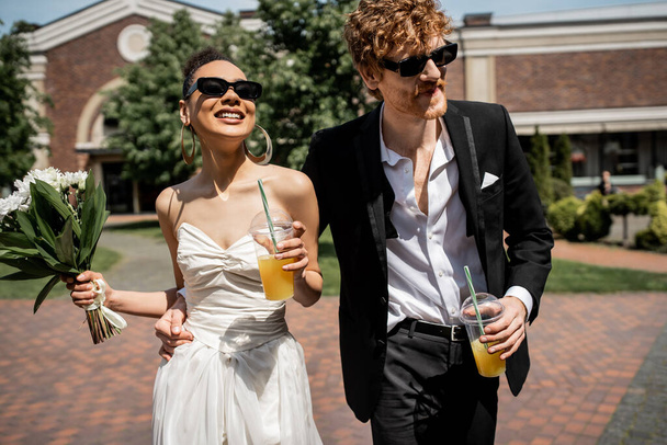 coppia interrazziale, occhiali da sole, abbigliamento da sposa, succo d'arancia, fiori, felicità, matrimonio all'aperto - Foto, immagini
