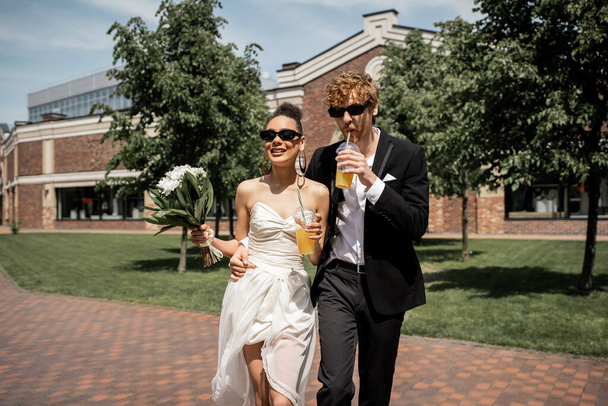 elegant newlyweds in sunglasses walking with orange juice and flowers in european city - Photo, Image