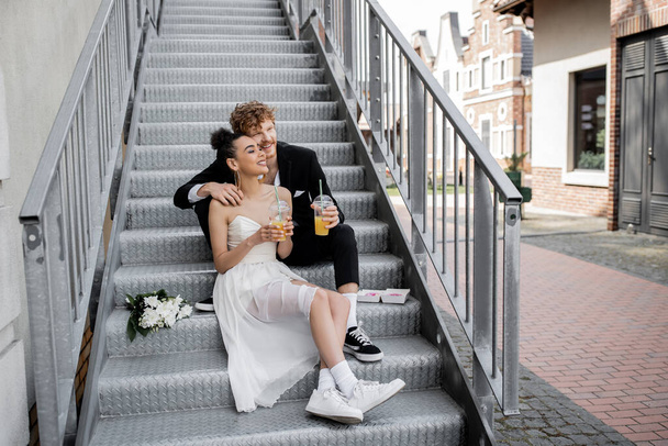 multiethnic newlyweds with orange juice sitting on stairs and looking away, wedding in city - Foto, immagini
