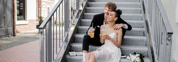 mariage dans la rue, heureux couple interracial assis sur les escaliers avec du jus d'orange, bannière - Photo, image